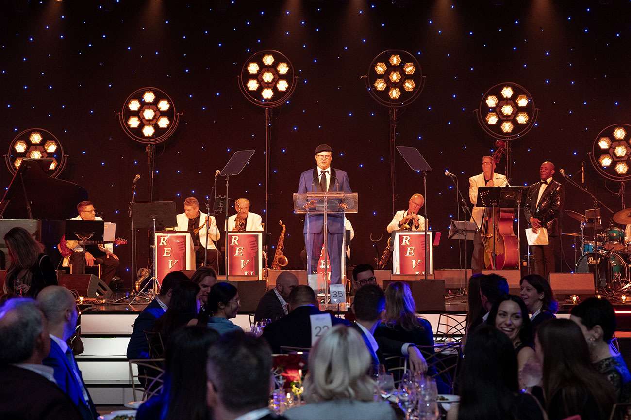 Man at podium on stage with musicians and MC in behind, speaking to room of dining attendees
